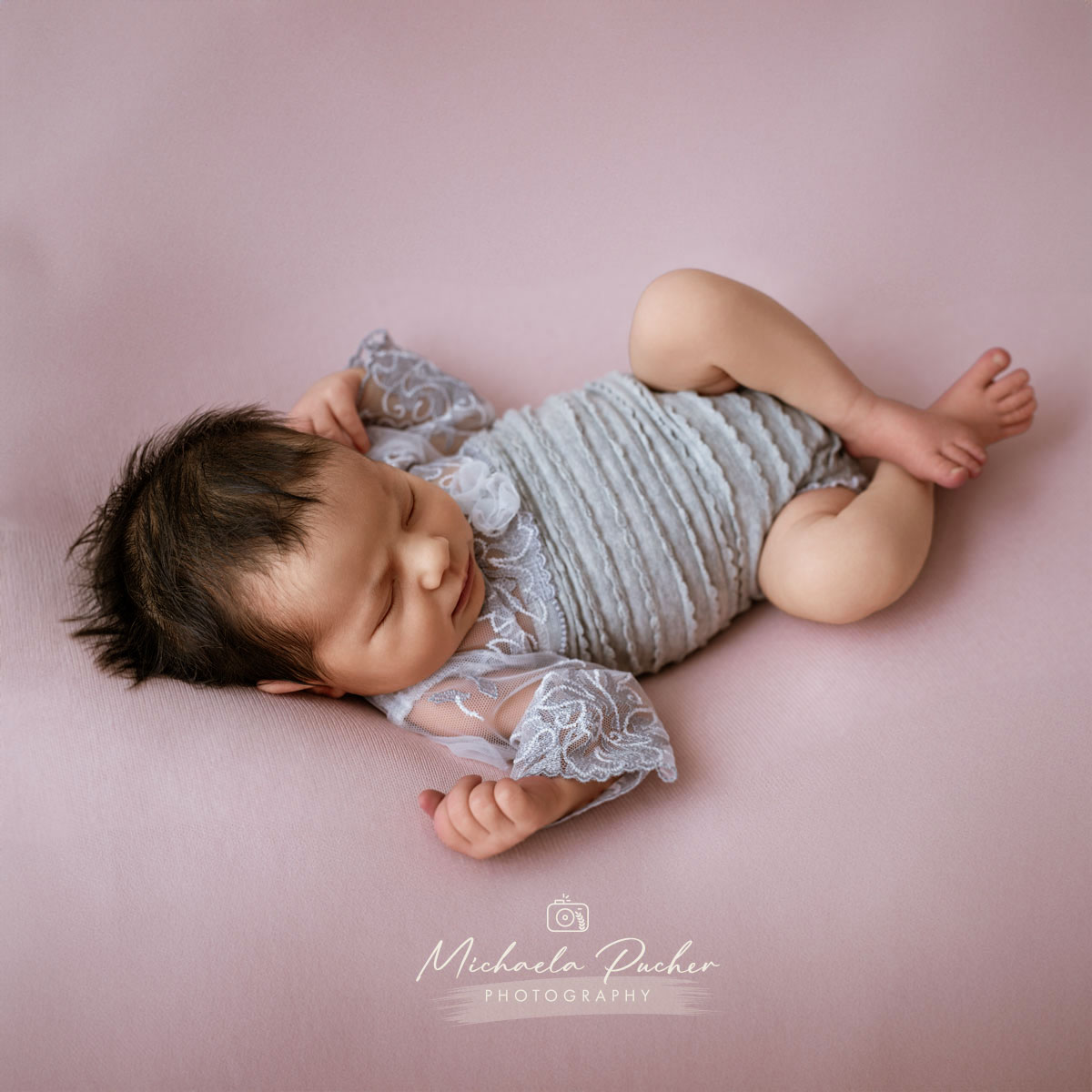 Schlafendes Neugeborenes in einem hellgrauen Spitzenbody, liegend auf einer rosafarbenen Unterlage, mit angewinkelten Beinchen und entspanntem Gesicht.